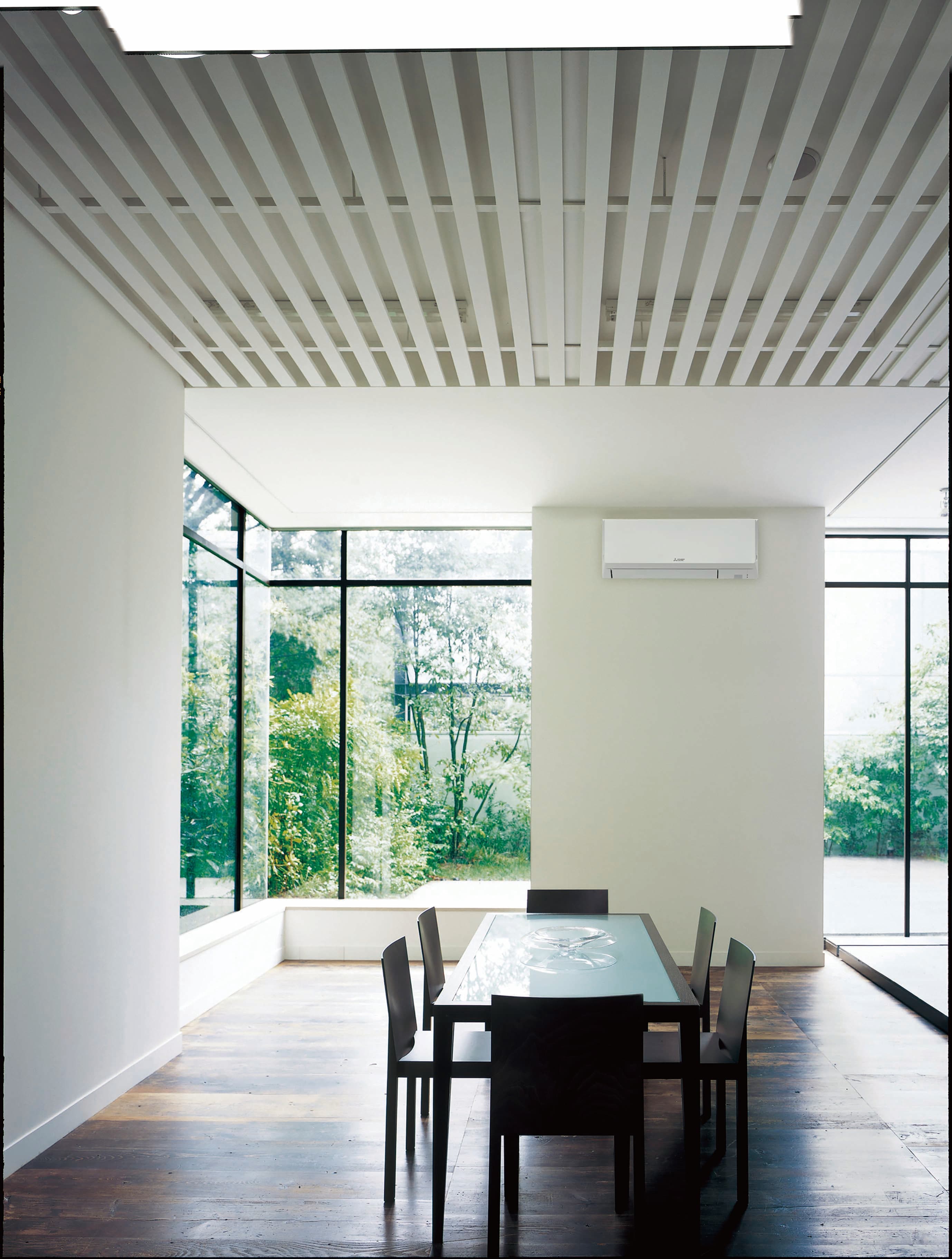 White wall-mounted heat pump above a dining table in a bright room with large windows.